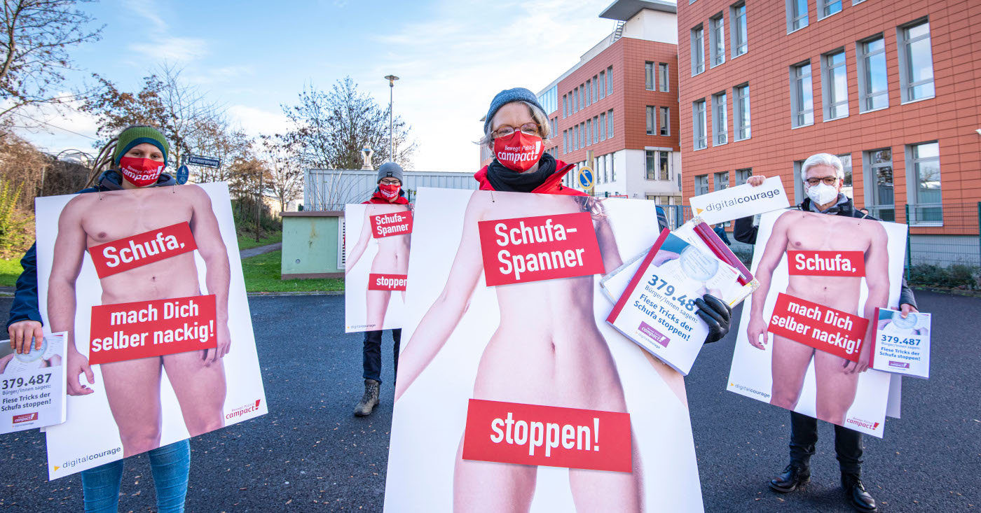Drei Personen halten Schilder mit nackten Menschen vor ihren Körpern. Auf den Schildern steht die Aufschrift "Schufa-Spanner stoppen"