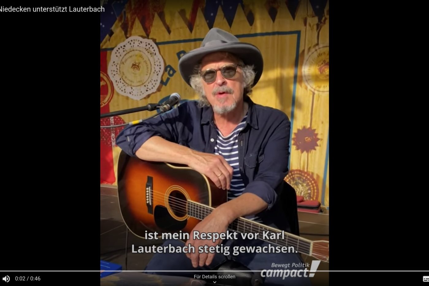 Screenshot aus dem Video "Wolfgang Niedecken unterstützt Lauerbach". Wolfgang Niedecken sitzt mit einer Gitarre auf dem Schoß vor einem Mikrofon und spricht in die Kamera