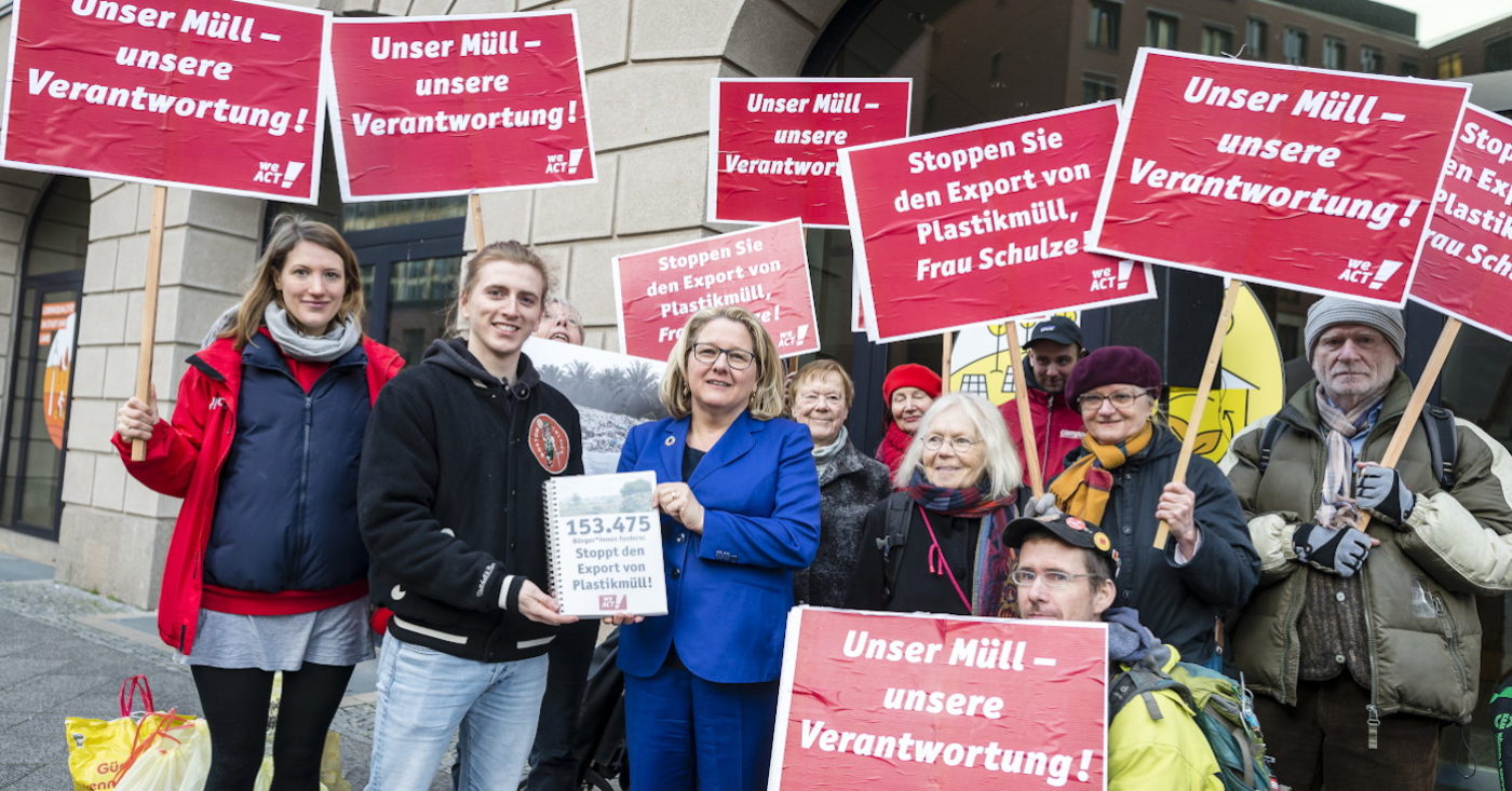 Max Hoffschmidt übergibt die Petition "Stoppt den Export von Plastikmüll!" an Umweltministerin Svenja Schulze. Im Hintergrund halten Demonstant*innen rote Schilder gegen den Export von Plastikmüll.