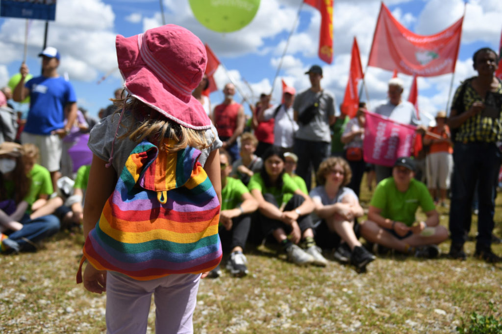 Ein Mädchen mit Regenbogenrucksack steht vor protestierenden Menschen mit Schildern.