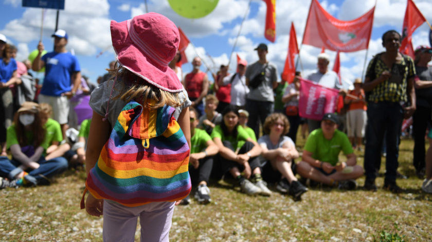 Ein Mädchen mit Regenbogenrucksack steht vor protestierenden Menschen mit Schildern.