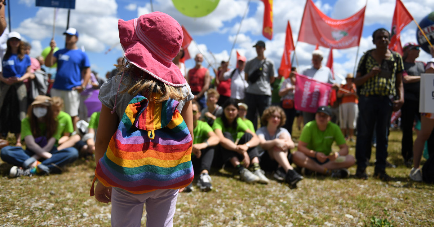 Ein Mädchen mit Regenbogenrucksack steht vor protestierenden Menschen mit Schildern.