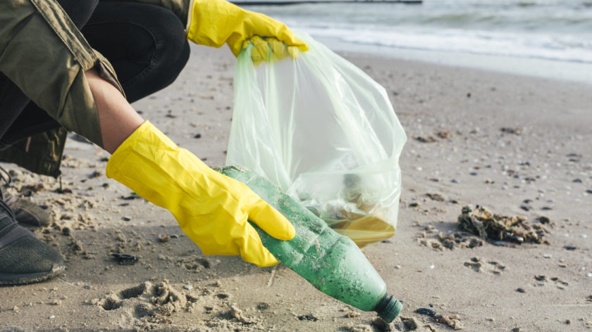 Zu sehen ist eine Person, die gelbe Gummihandschuhe trägt und am Strand eine Plastikflasche aufsammelt. In der anderen Hand hält sie eine Tüte, in der bereits weiterer Müll ist.