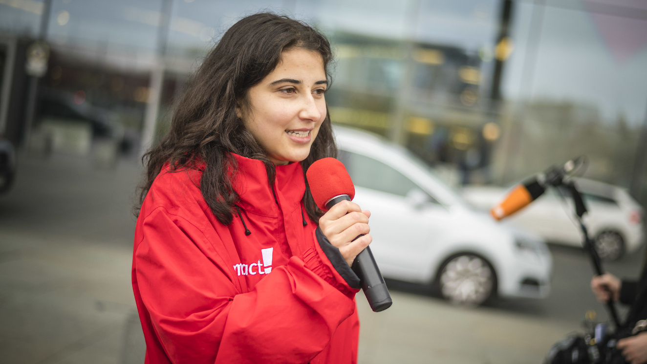 Das Foto zeigt Campact-Campaignerin Friederike Gravenhorst. Sie hat eine rote Jacke an, auf der links das Campact-Logo in Weiß zu sehen ist. Sie hält ein Mikorfon in der Hand, in das sie hineinspricht. Das Foto wurde bei einer Aktion von Campact aufgenommen.