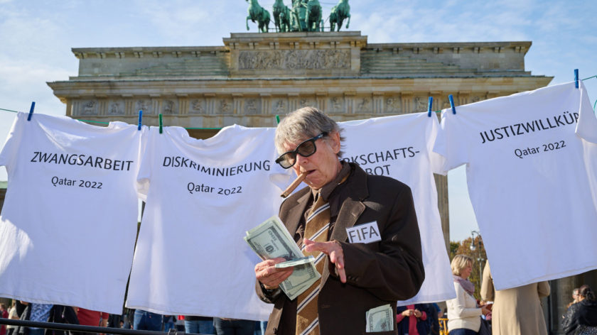 Das Foto zeigt eine Protestaktion vor dem Brandenburger Tor in Berlin. Eine grauhaarige Person steht im Vordergrund und hat viele Geldscheine in der Hand. Auf ihrer Jacke ist ein Aufkleber, auf dem FIFA steht. Im Hintergrund ist eine Wäscheleine gespannt, auf der weiße T-Shirts hängen. Darauf stehen die Begriffe Diskriminierung, Justizwillkür und Zwangsarbeit.