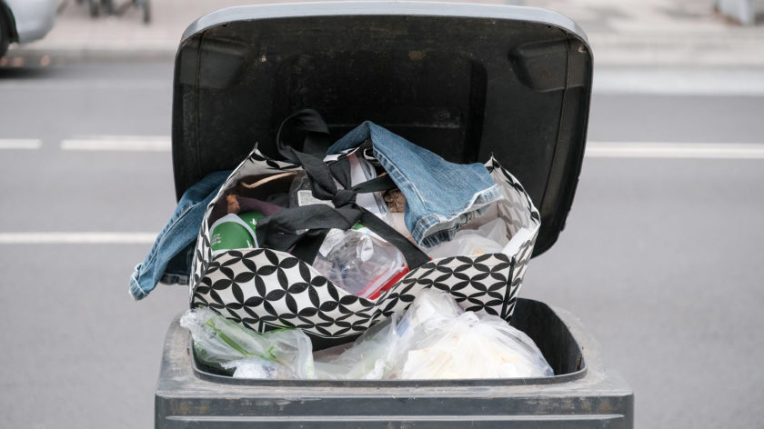 Das Foto zeigt eine geöffnete schwarze Mülltonne. Sie ist voll mit verschiedenen Plastiktüten und zugebundenen Beuteln. Oben auf liegt eine kaputte Jeanshose.