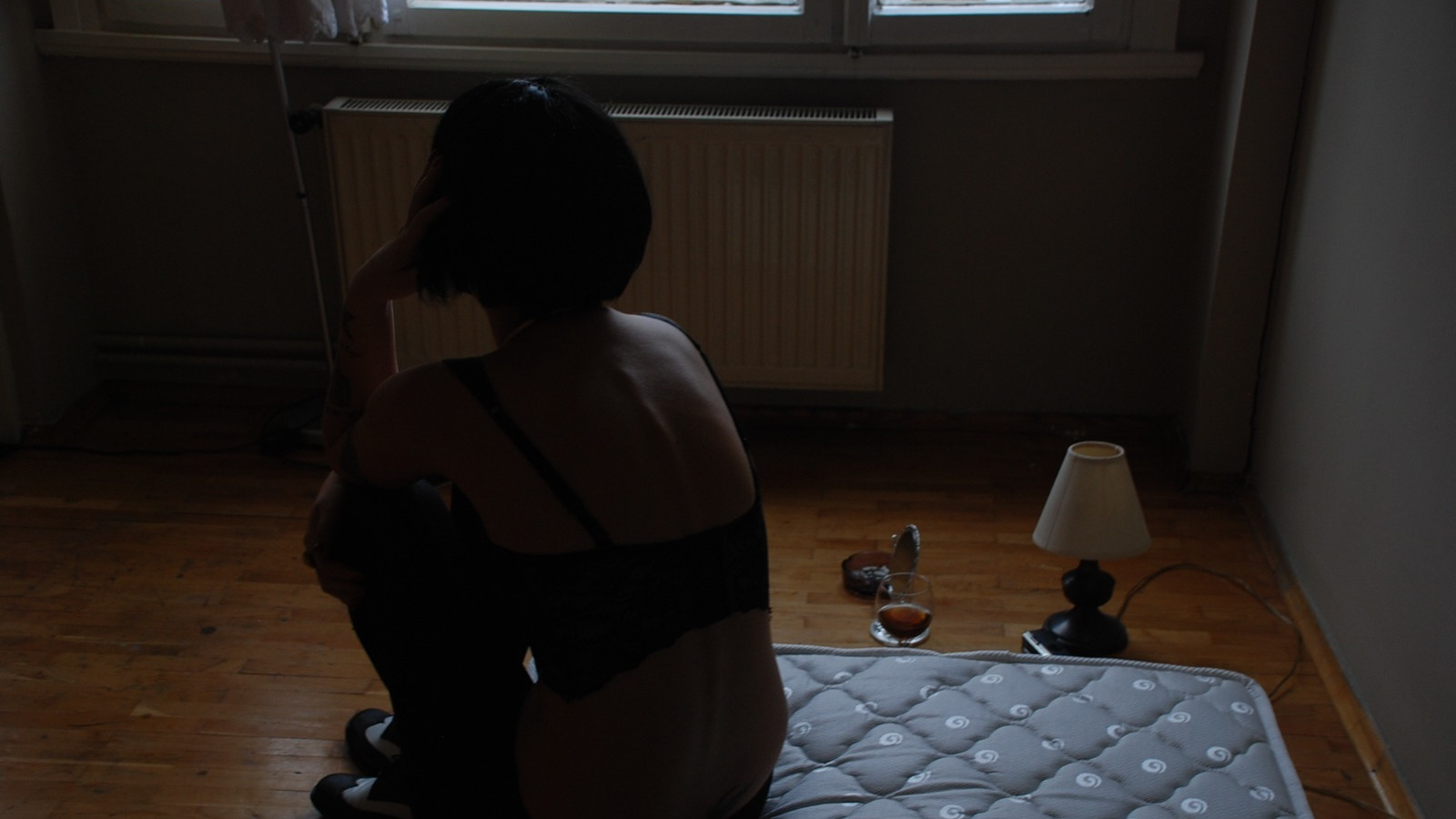 Das Foto von Nazife Behramoğlu (Istanbul 2011) zeigt eine Frau, die in einen schwarzen BH und eine schwarze Hose gekleidet auf einer Matratze sitzt, die auf dem Boden liegt. Die Frau hat kinnlange, schwarze Haare, sie sitzt mit dem Rücken zu uns. Neben ihr auf dem Boden stehen ein Glas , ein kleiner Spiegel, ein Aschenbecher und eine kleine Lampe.