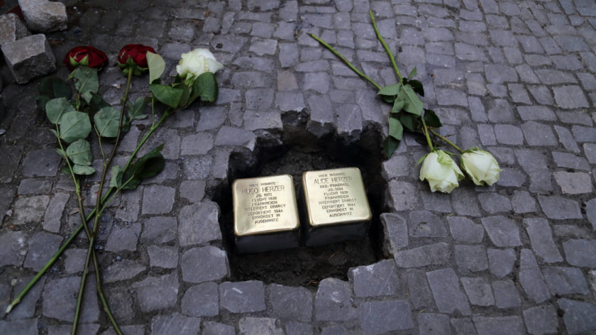 Das Foto zeigt eine Stolpersteinverlegung in Potsdam. Zwei Stolpersteine sind in den Boden eingelassen, aber noch nicht in den Bürgersteig eingeschlossen. Neben den Steinen liegen Rosen.