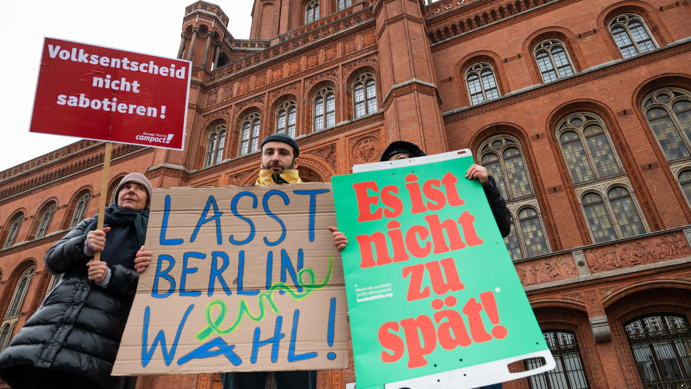 Drei Menschen mit verschiedenen bunten Schildern, auf denen Protestsprüche stehen, stehen vor einem großen, roten Backsteingebäude. Es ist das Rote Rathaus in Berlin.