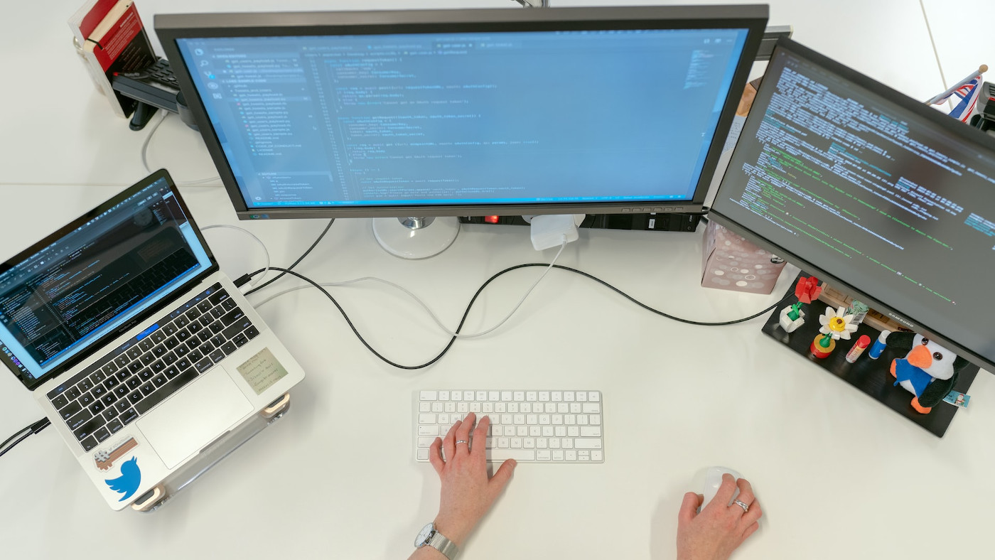 Das Foto zeigt einen Schreibtisch von oben. auf ihm stehen links ein aufgeklappter Laptop, in der Mitte und rechts steht jeweils ein Bildschirm. Auf allen Bildschirmen sind Code-Zeilen zu sehen. Am unteren Bildrand sieht man noch eine Tastatur und eine Computermaus, auf denen jeweils eine Hand liegt.
