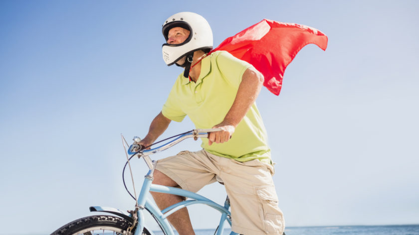 Das Foto zeigt einen Mann, der auf einem blauen Fahrrad sitzt. Er trägt eine helle Short-Hose, ein gelbes T-Shirt, einen Mofa-Helm und ein rotes Superhelden-Cape.
