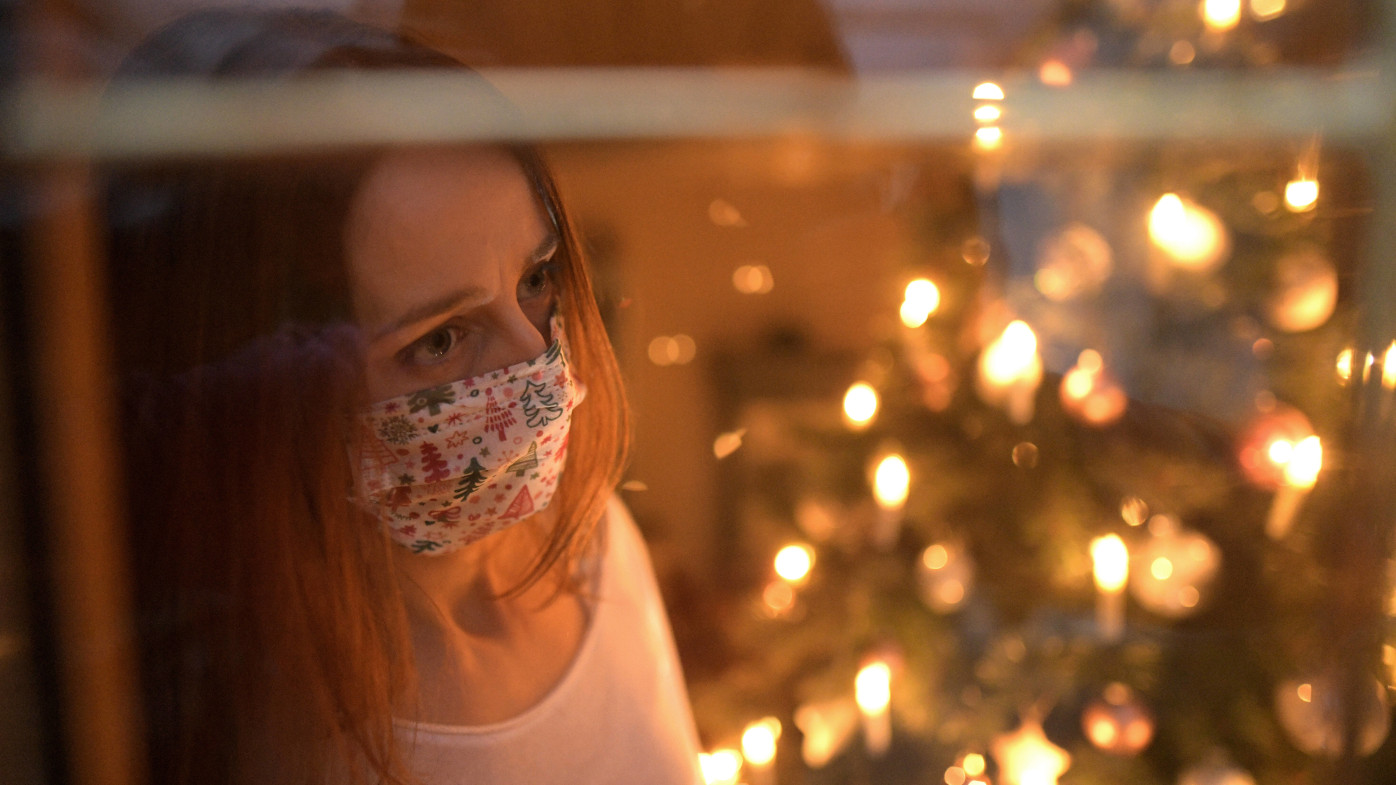 Das Foto zeigt eine Frau in einem weihnachtlich geschmückten Raum vor einem Weihnachtsbaum. Sie trägt einen Mund-Nase-Schutz und schaut aus dem Fenster.