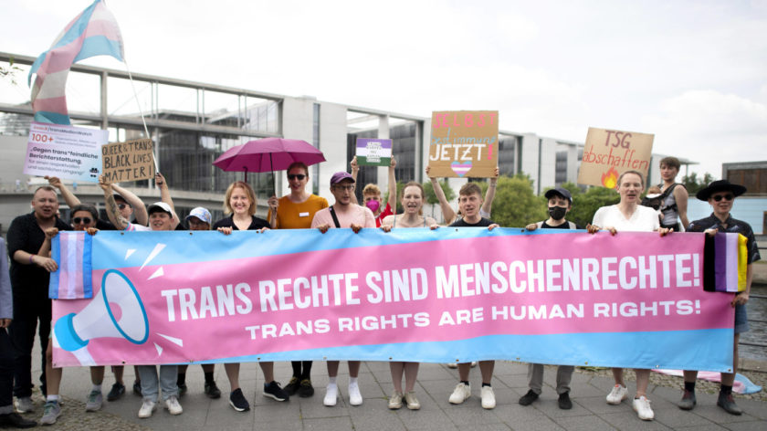 Das Foto zeigt mehrere Personen, die ein blau-rosa-farbenes Banner mit einem weißen Schriftzug darauf hochhalten.