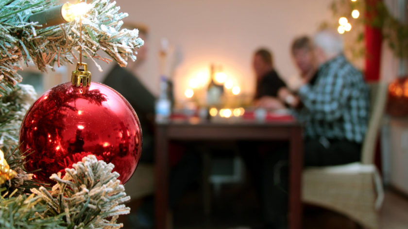 Eine Familie sitzt an Heiligabend gemeinsam am Tisch und geniesst das Weihnachtsessen. Im Vordergrund ist eine rote Weihnachtskugel und ein Zweig von einem Weihnachtsbaum zu sehen.
