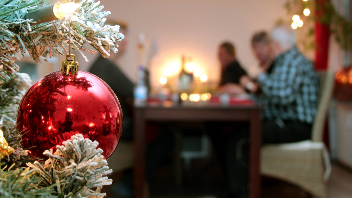 Eine Familie sitzt an Heiligabend gemeinsam am Tisch und geniesst das Weihnachtsessen. Im Vordergrund ist eine rote Weihnachtskugel und ein Zweig von einem Weihnachtsbaum zu sehen.