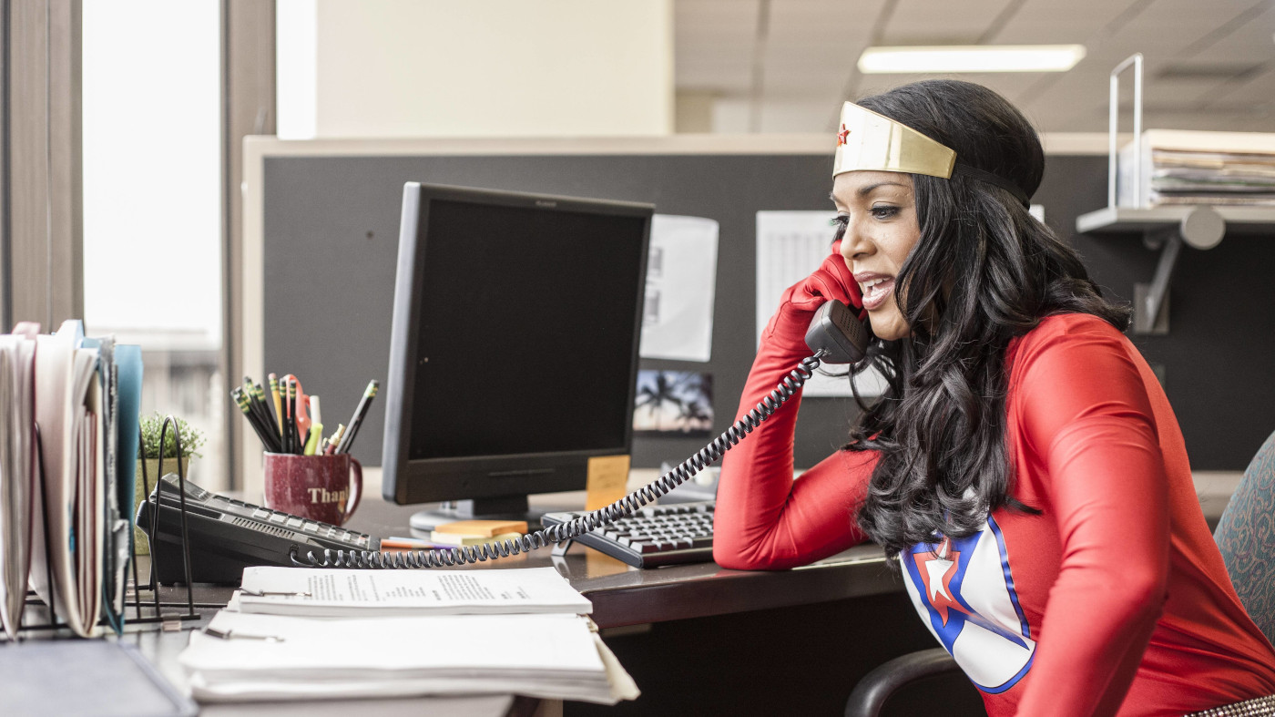 Eine Frau im Superheldinnen-Kostüm sitzt am einem Schreibtisch, auf dem viele Büro-Unterlagen liegen. Sie hat einen Telefonhörer in der Hand, der mit dem Telefon auf dem Tisch verbunden ist. So könnte die Arbeit für eine zukünftige Familienministerin sinnbildlich aussehen.