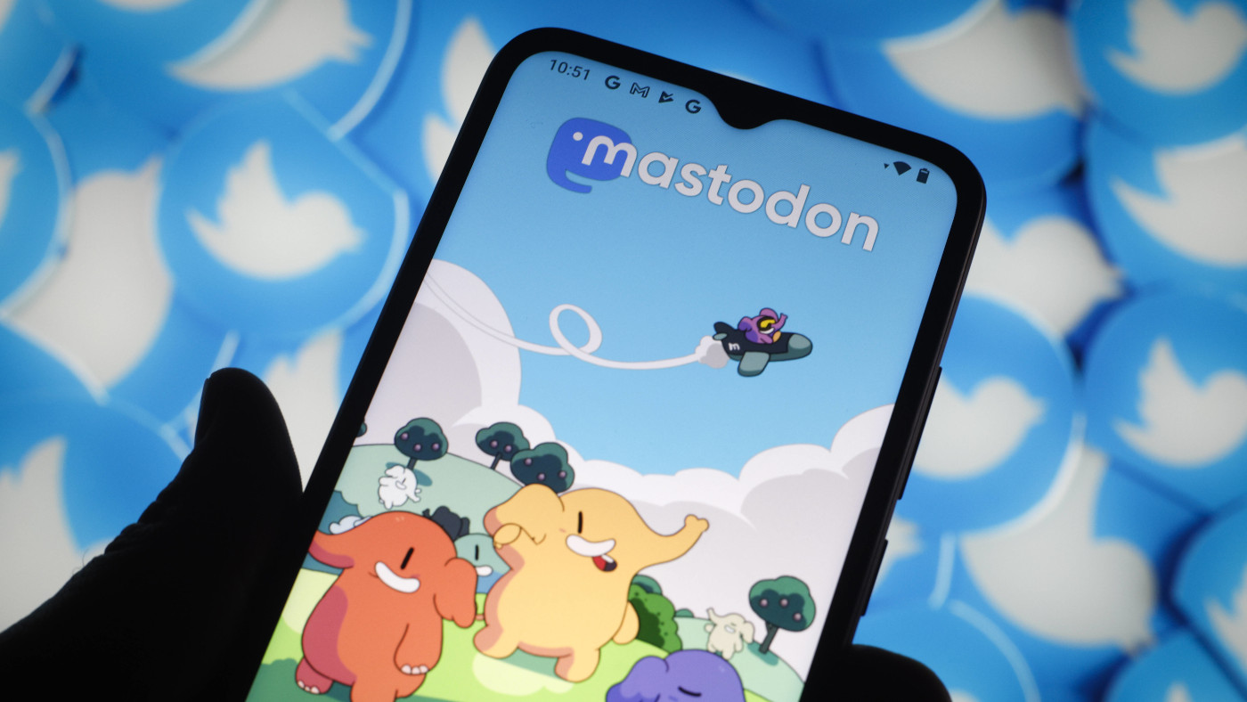 Zu sehen ist im Vordergrund ein Smartphone, auf dem der Startbildschirm des Netzwerks Mastodon zu sehen ist. Im Hintergrund sind unscharf mehrere Ausführungen des Logos von Twitter zu erkennen.