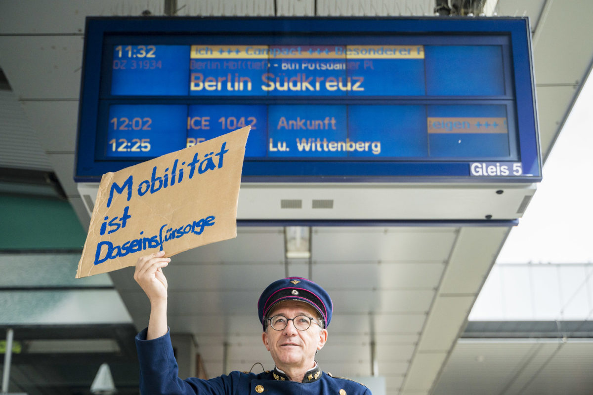 Das Foto zeigt einen als Schaffner gekleideten Mann, der ein Schild hochhält, auf dem steht "Mobilität ist Daseinsfürsorge". Hinter ihm ist eine Anzeigetafel von einem Bahnhof zu sehen.