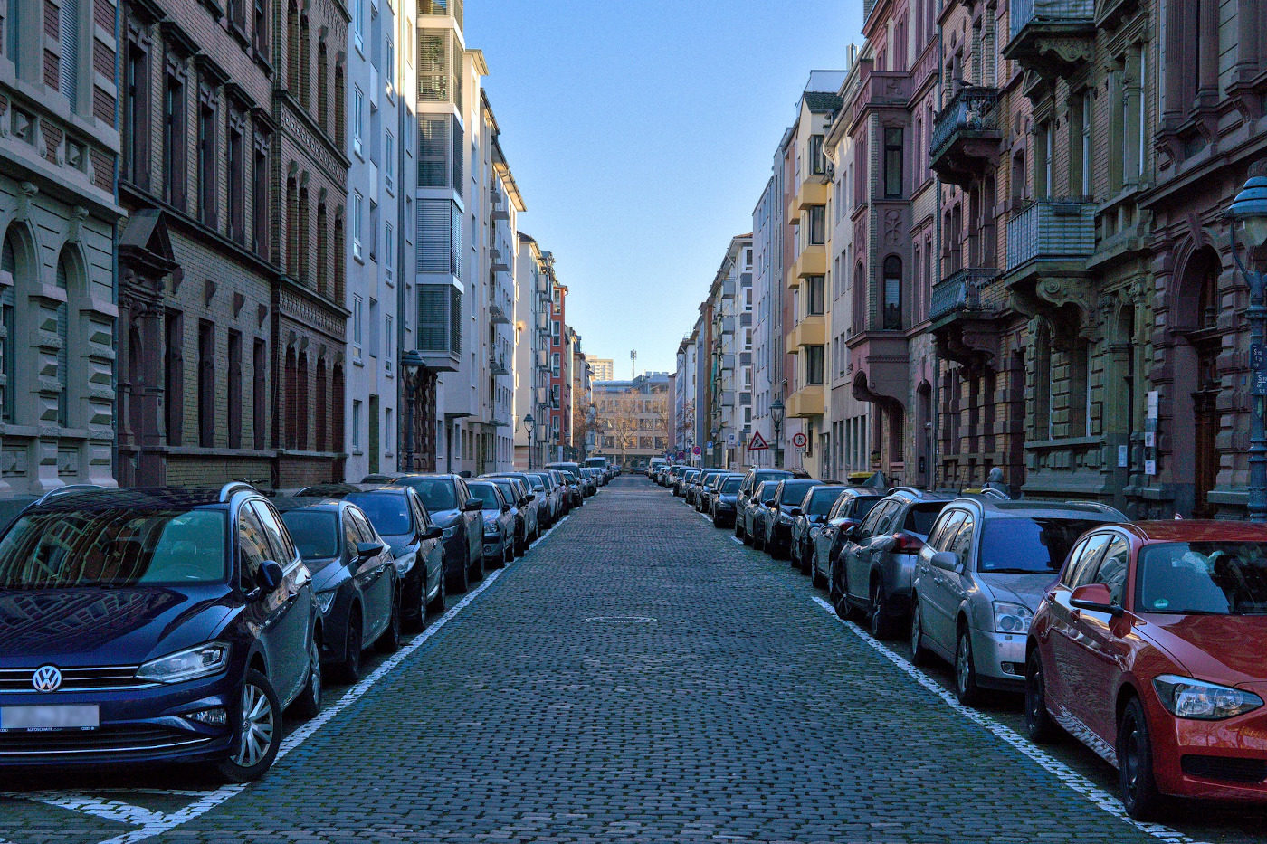 Das Foto zeigt eine Straße mit Kopfsteinpflaster. Rechts und links stehen dicht an dicht Häuser. Auf beiden Seiten parken viele Autos am Straßenrand.