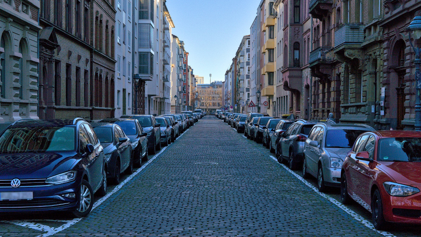 Das Foto zeigt eine Straße mit Kopfsteinpflaster. Rechts und links stehen dicht an dicht Häuser. Auf beiden Seiten parken viele Autos am Straßenrand.