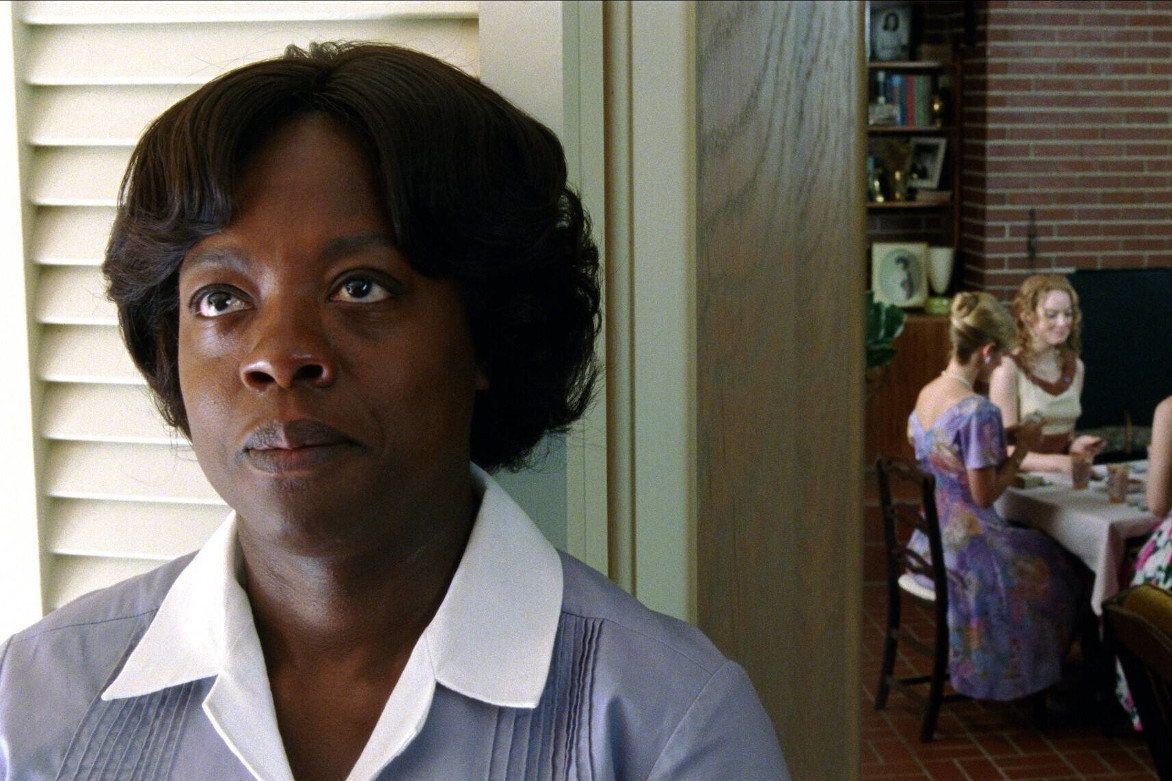Das Bild zeigt ein Standbild aus dem Film "The Help" (deutscher Titel: "Gute Geister"). Zu sehen ist Aibileen (gespielt von Viola Davis) im Vordergrund, sie steht am Rahmen einer offenen Tür. Im Hintergrund sind vier weiße Frauen in bunten Kleidern zusehen, die um einen Tisch sitzen.