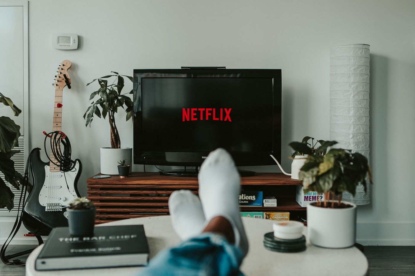 Eine Person sitzt auf einem Sofa, das Foto ist aus ihrer Perspektive aufgenommen, ihre Füße liegen auf einem Tisch. Sie schaut auf einen Fernseher, auf dem das Netflix-Logo zu sehen ist.