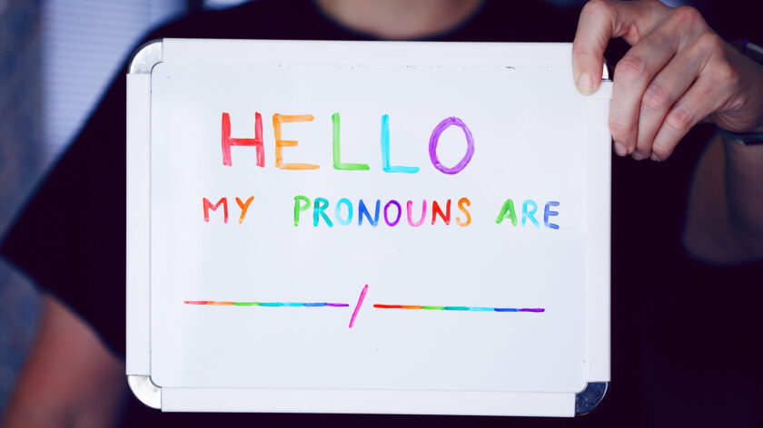 Ein Mensch hält ein Schild hoch auf dem steht: Hallo, meine Pronomen sind ....