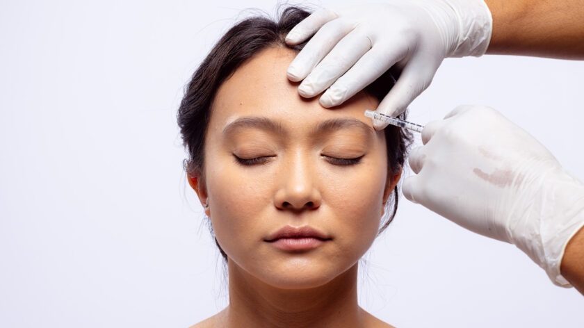 Hände einer Kosmetikerin in weißen OP-Handschuhen spritzen Botox in die Stirn einer asiatischen Frau