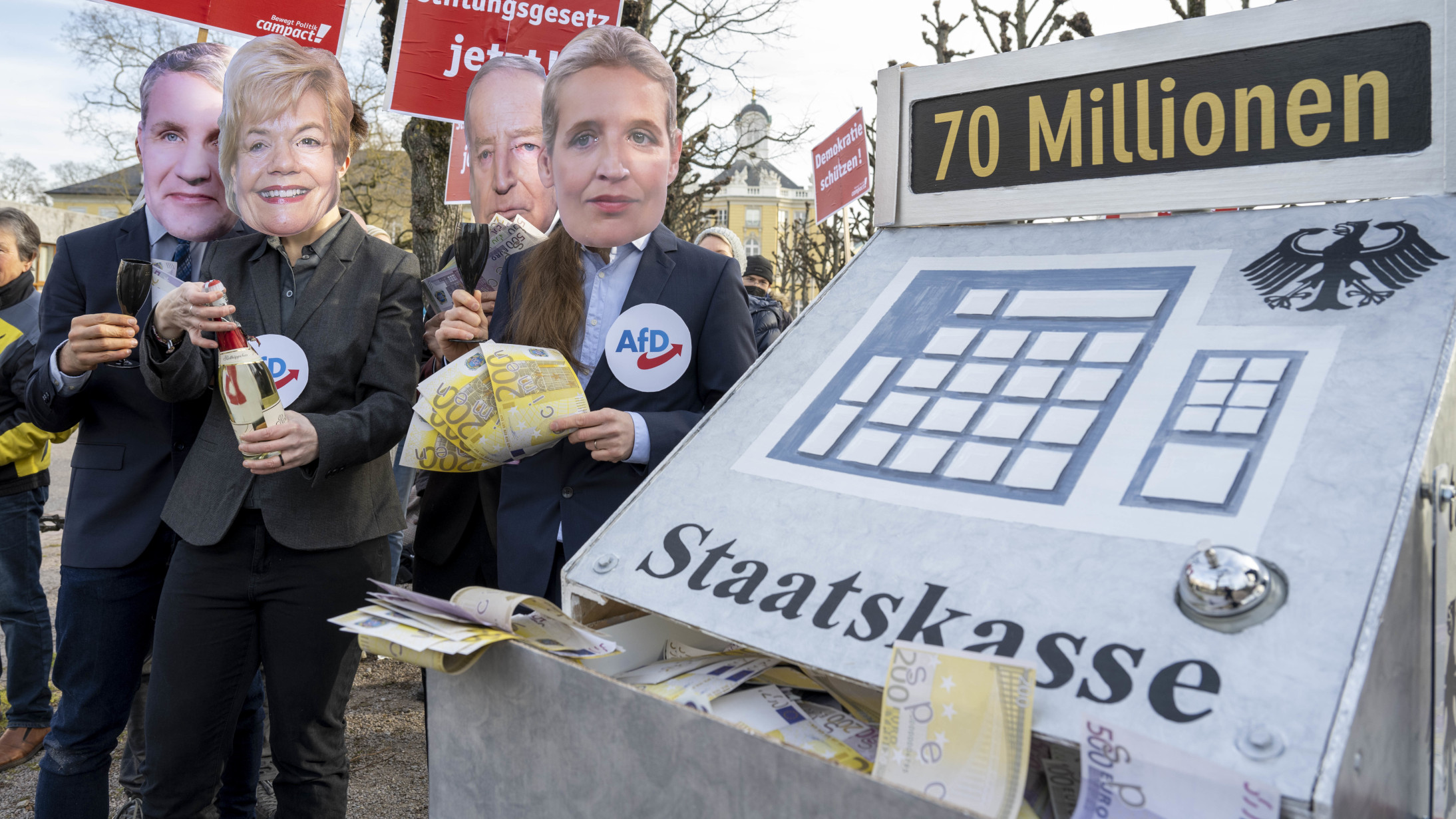 Vier Darsteller mit Masken von Personen aus dem Umfeld der AfD stehen neben einer stilisierten Staatskasse, an der sie sich bedienen. Das Foto ist im Rahmen einer Protest-Aktion vor dem Bundesverfassungsgericht in Karlsruhe im Februar 2023 entstanden.