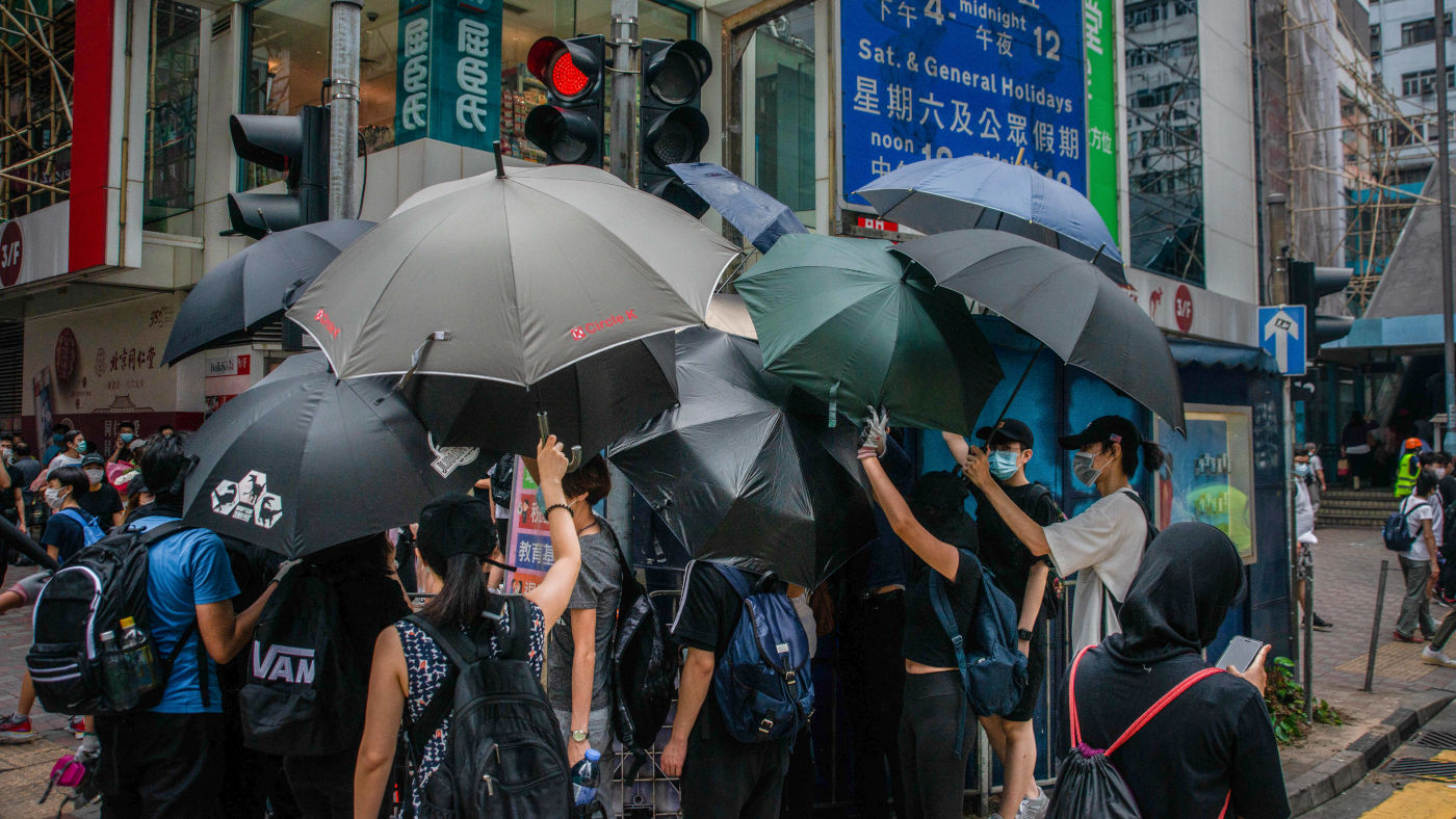 Das Foto zeigt Menschen in Hong Kong, die an einer Straßenkreuzung stehen. Sie halten viele Regenschirme in die Luft und seitlich um sich herum. Damit schirmen sie sich vor dem Blick von Überwachungskameras ab.