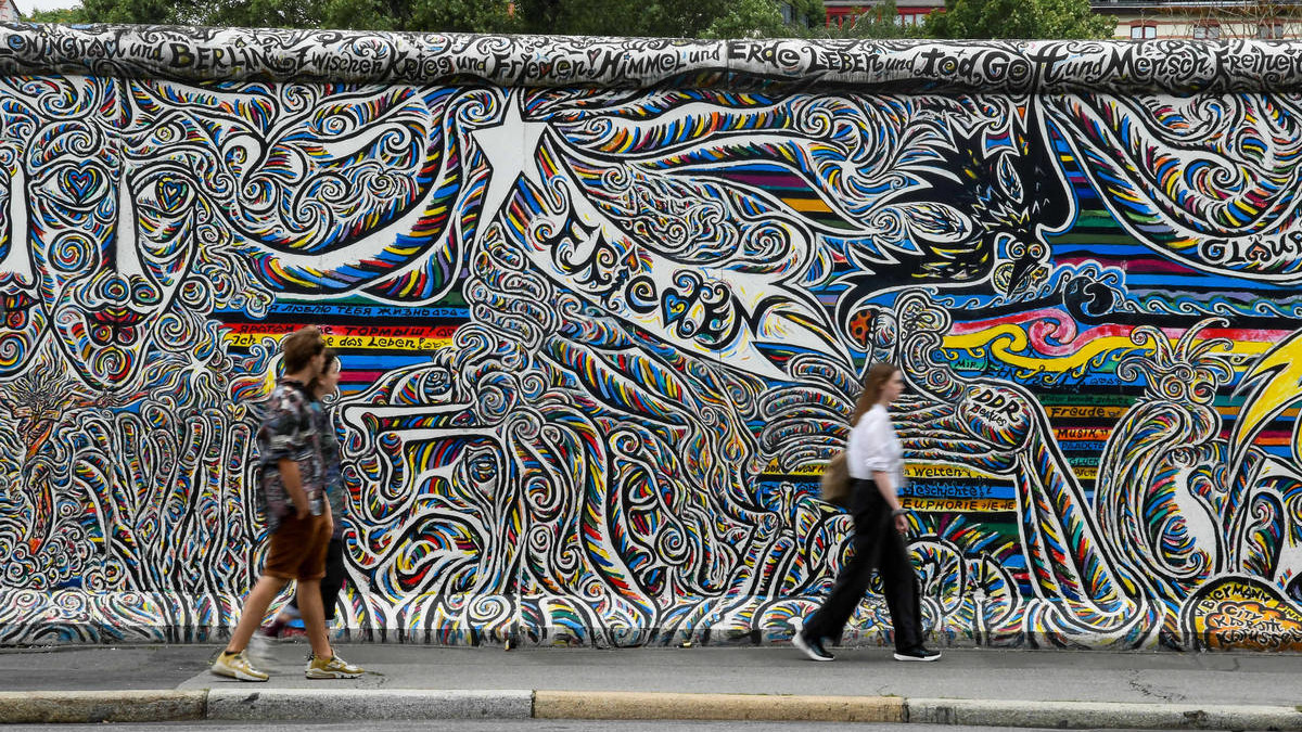 Das Foto zeigt ein Stück der Mauer an der East Side Gallery in Berlin in der Frontalaufnahme. Es sind viele bunte Linien und Symbole zu erkennen. Vereinzelt kann man Wörter lesen: Frieden, Berlin, Sehnsucht. Im Vordergrund gehen drei Personen von links nach rechts durchs Bild an der Mauer vorbei.