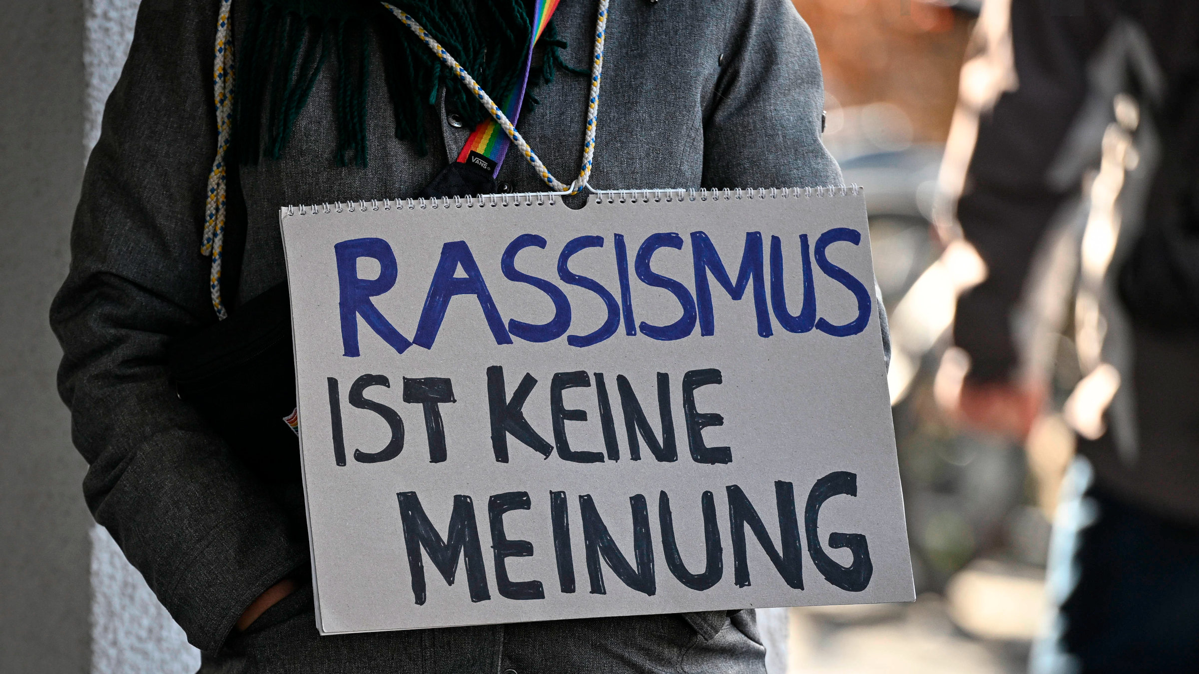 Eine Person auf einer Demo hat ein Schild um den Hals gehängt. Darauf steht: Rassismus ist keine Meinung.