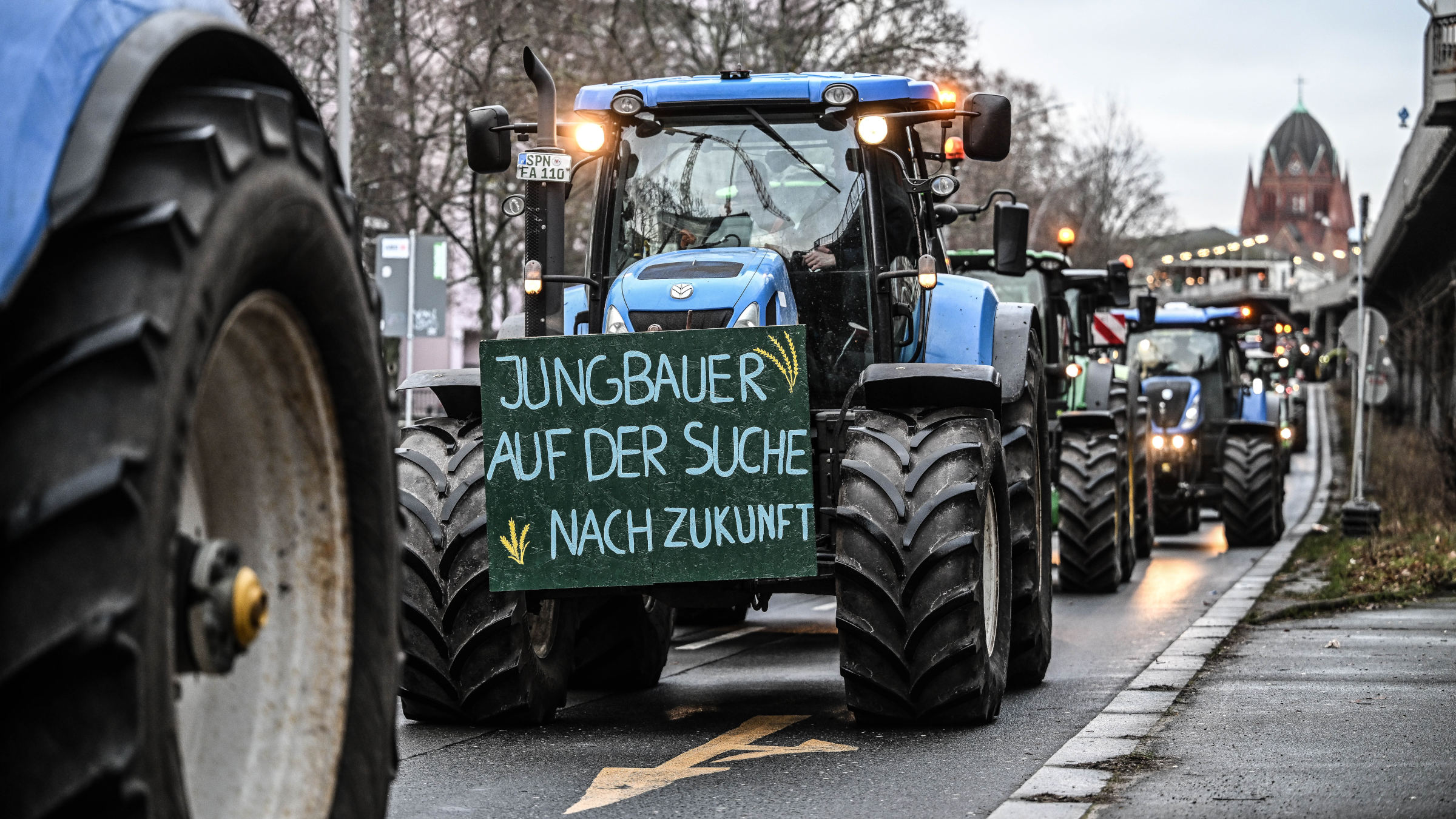 Landwirt*innen protestieren vor dem Brandenburger Tor. An einem blauen Traktor hängt vorne ein Schild: Jungbauer auf der Suche nach Zukunft.