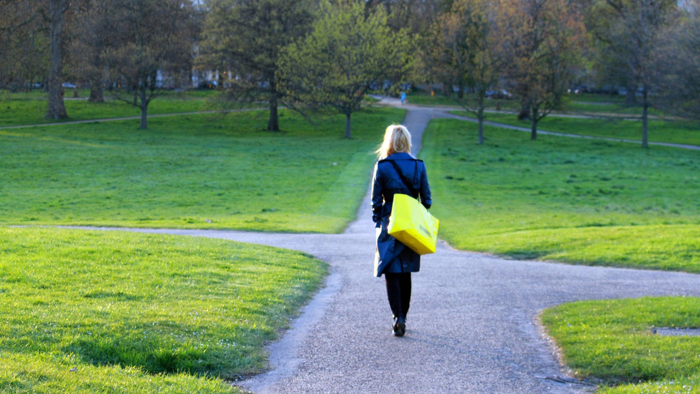 Das Foto zeigt eine Frau von hinten, die einen Weg in einem Park entlanggeht. Sie hat blonde Haare, die sie offen trägt. Sie trägt außerdem einen langen blauen Mantel und eine viereckige gelbe Tasche.