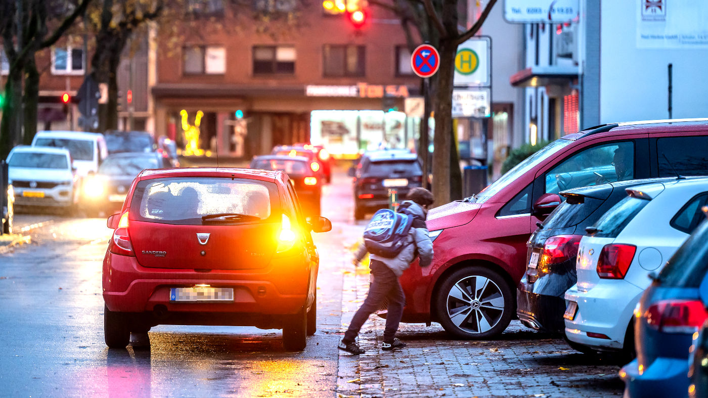 Mehrere Autos ("Elterntaxis") halten auf einer Straße vor einer Schule in NRW. Zentral steht en kleines rotes Auto, das den rechten Blinker gesetzt hat. Davon weg läuft ein kleiner Junge mit Schulranzen zwischen parkende Autos.