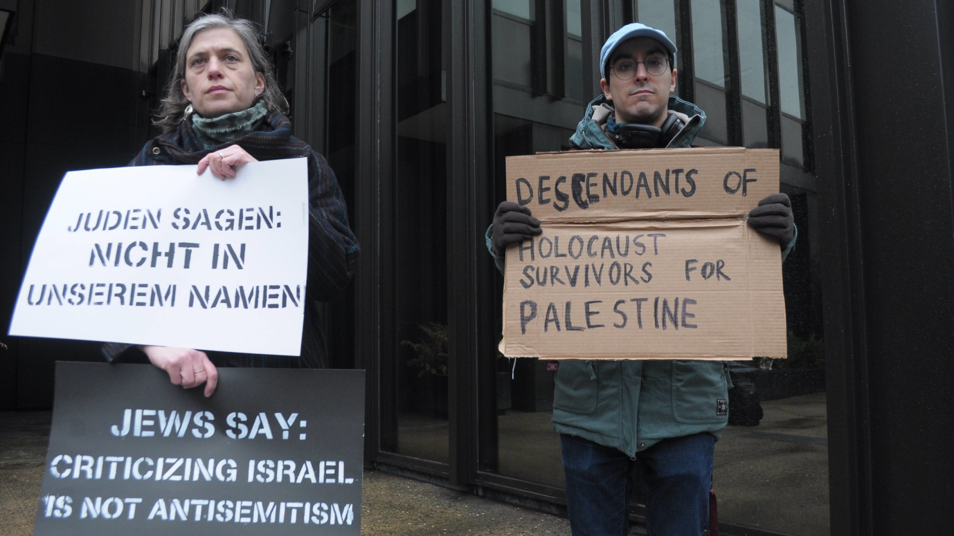 Zwei Personen stehen draußen vor einem Bürogebäude und halten Schilder. Auf den Schildern der linken Person steht: "Juden sagen: Nicht in unserem Namen" und "Jews say: Criticizing Israel is not Antisemitism." Auf dem Schild der rechten Person steht: "Descendants of Holocaust Survivors for Palestine".