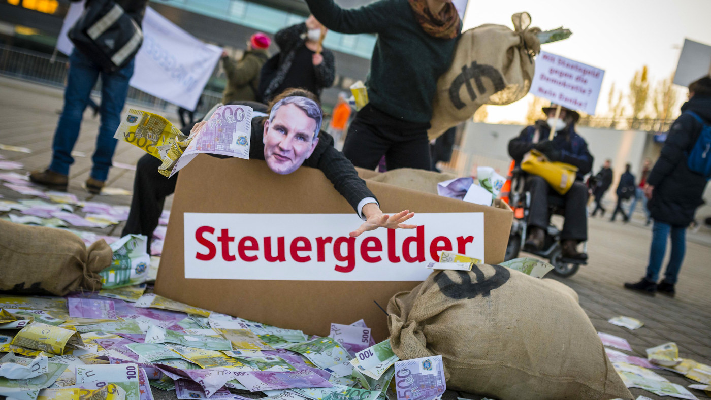Darsteller mimen AfD-Politiker, die in Geld baden. Vor einem Schauspieler, der Björn Höcke darstellt, liegt viel Spielgeld und ein Karton, auf dem groß "Steuergelder" drauf steht.