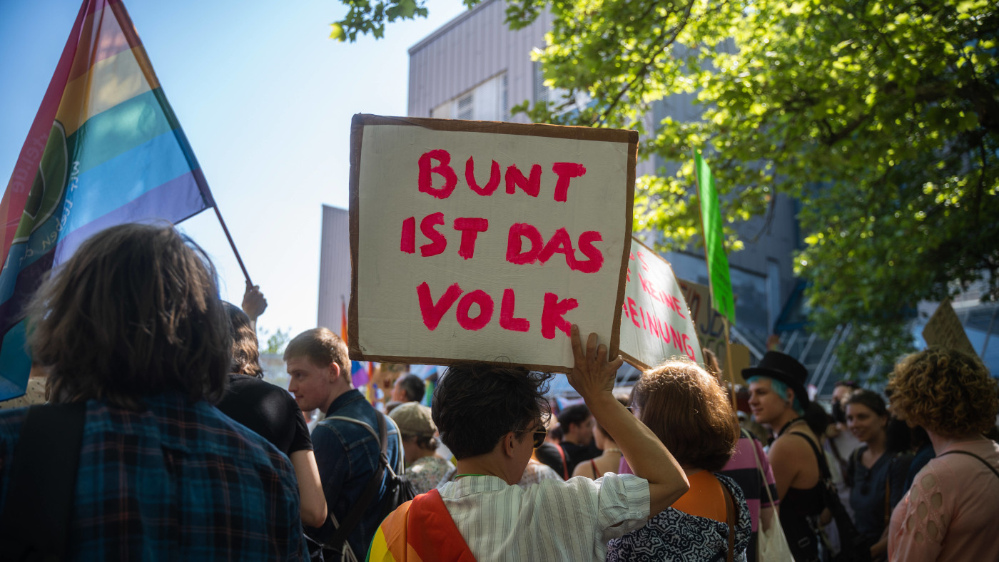 Das foto zeigt mehrere Personen von hinten mit Protestschildern. Das Schild in der Mitte zeigt den Text "Bunt ist das Volk".