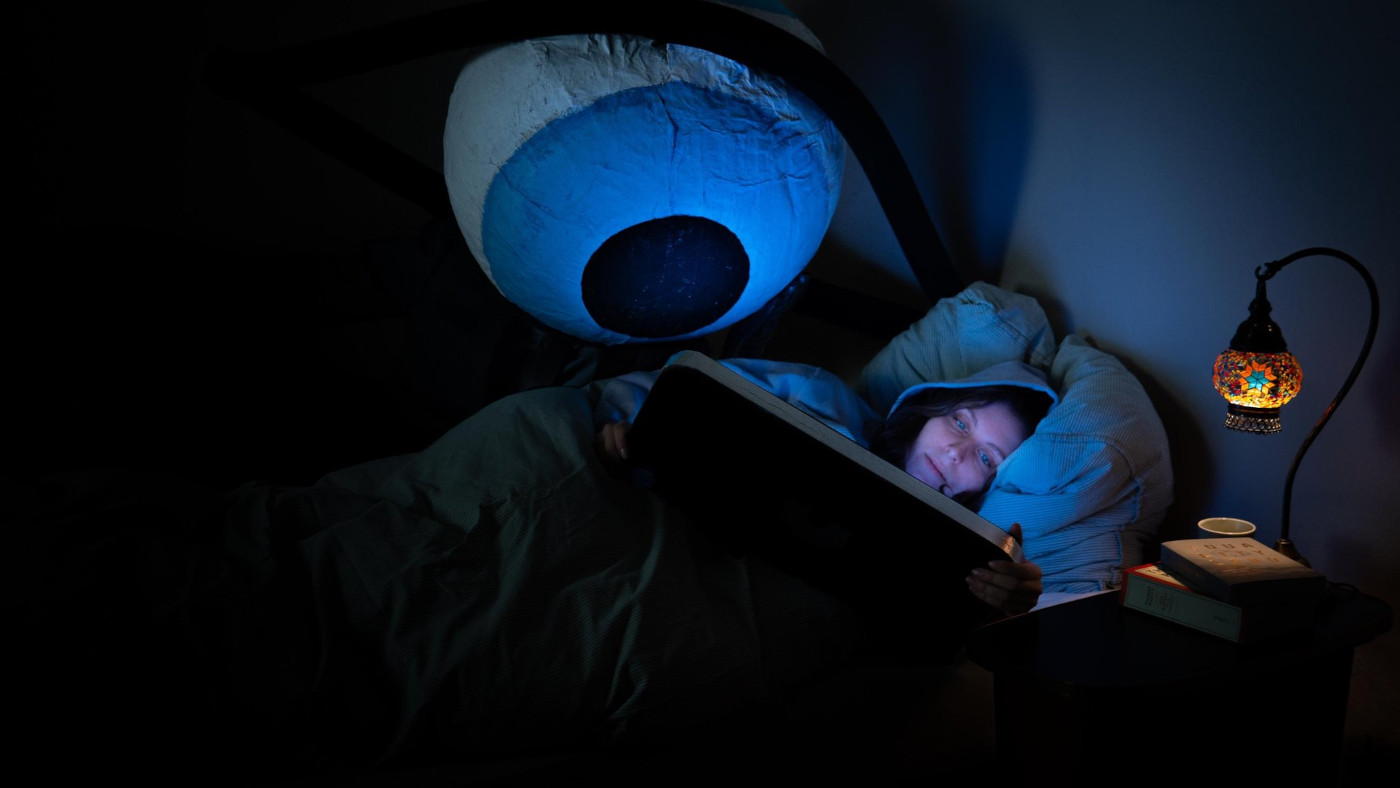 Das Foto zeigt eine Frau, die in einem dunklen Raum im Bett auf ihrer Seite liegt. In den Händen hat sie ein übergroßes Smartphone, von dessen Bildschirm ein blaues Licht ausgeht. Hinter ihr neben dem Bett steht ein großes, stilisiertes Auge, das mit auf den Bildschirm schaut.