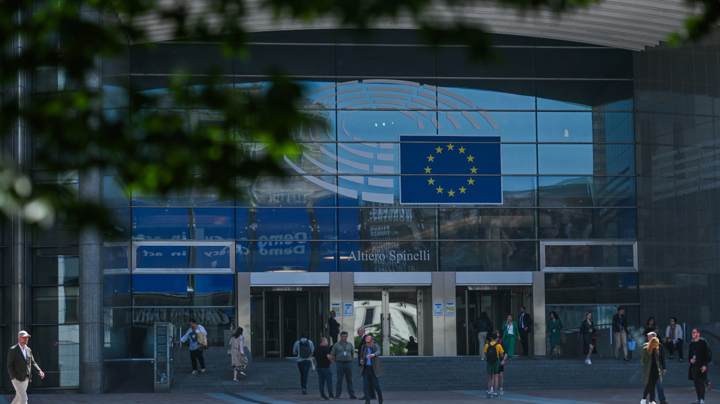 Das Foto zeigt den Eingang des EU-Parlaments in Brüssel in einer Großaufnahme. Vor dem Eingang stehen einige Menschen in kleinen Gruppen. Das Foto wird am oberen Rand von unscharfen Blättern im Vordergrund eingerahmt.