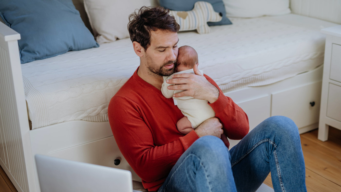 Das Foto zeigt einen Vater, das ein neugeborenes Baby im Arm hält. Er sitzt mit dem Rücken zu einem Sofabett. Neben ihm steht ein offener Laptop.