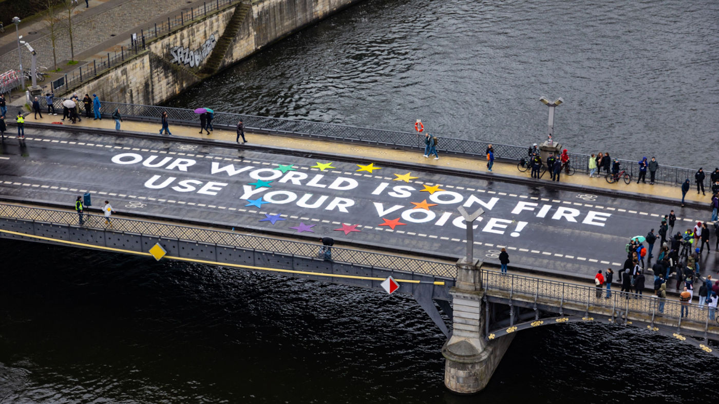 Personen stehen auf einer Brücke neben einem Schriftzug, der auf den Asphalst geschrieben ist: "Our world is on fire, use your voice!"