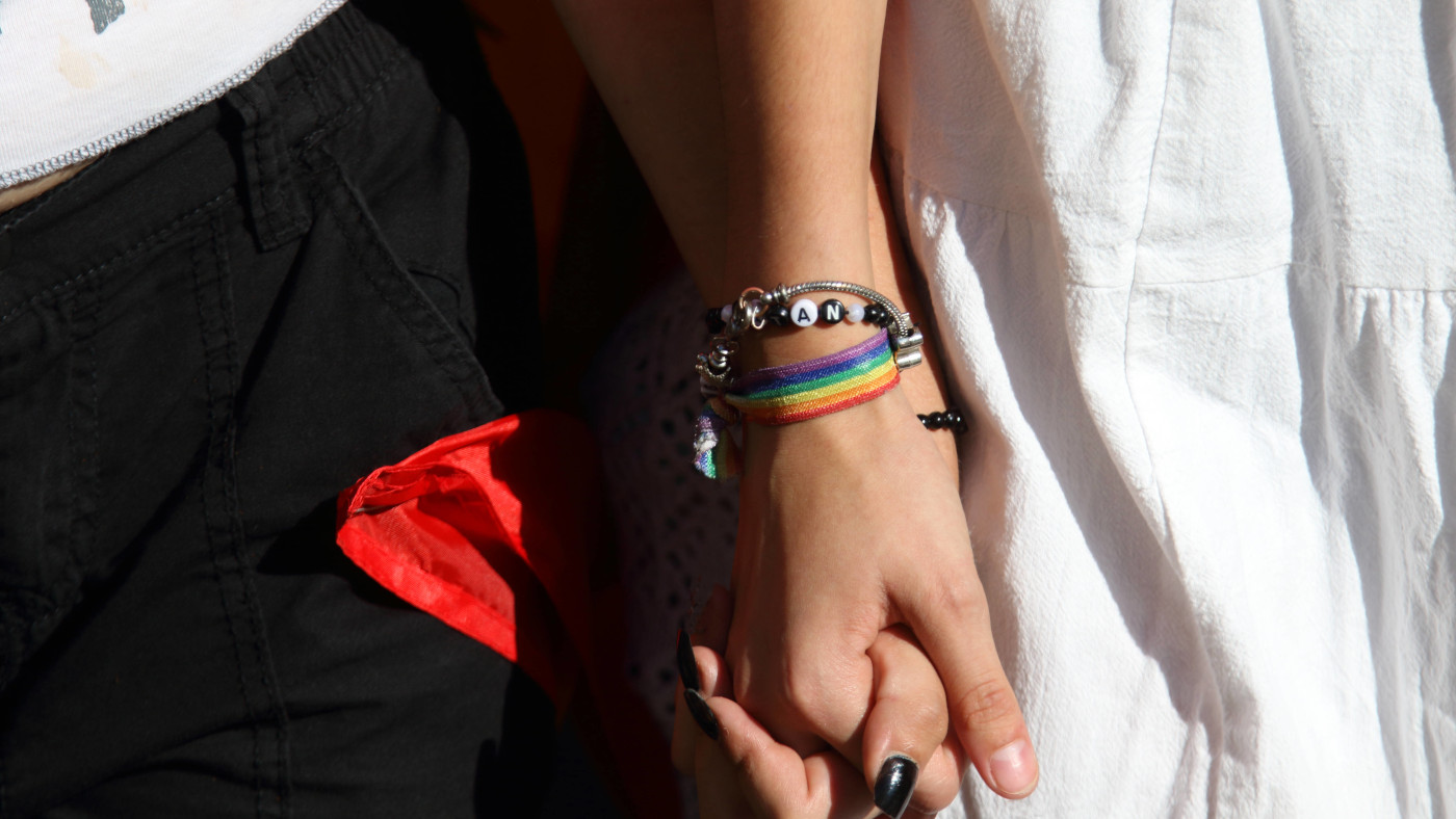 Demoteilnehmer*innen halten sich bei einer Pride-Demo an der Hand. Eine Person trägt ein Armband in Regenbogenfarben, die andere eine rote Schleife.