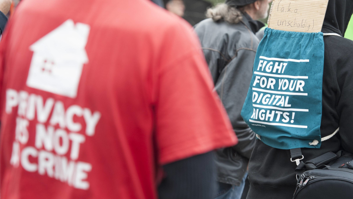 Der Oberkörper einer Person ist im linken vorderen Bildausschnitt zu sehen. Sie trägt ein rotes T-Shirt mit der aufschrift "Privacy is not a crime". In der rechten Bildhälfte sieht man den Rücken einer Person, die einen Zugbeutel trägt. Er hat die Aufschrift: "Fight for your digital rights".