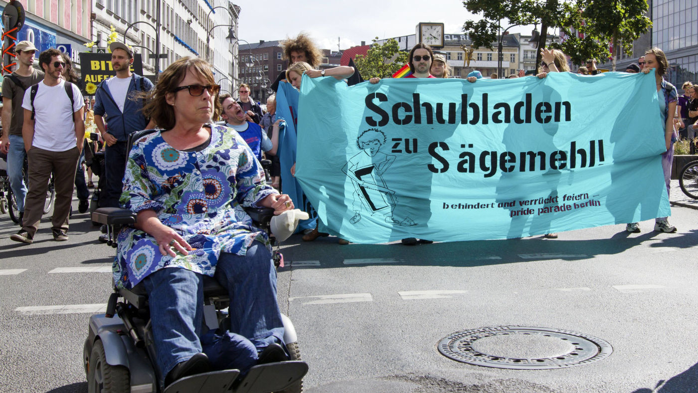 Eine Teilnehmerin im Rollstuhl und andere Menschen mit Plakaten und Bannern bei der Disability Pride Parade 2014 in Berlin. Auf einem großen Banner im Hintergrund steht "Schubladen zu Sägemehl!"