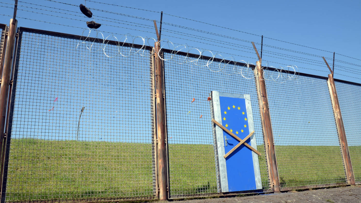 Symbolbild: Ein hoher Zaun mit Stacheldraht und einer verbarrikadierten Tür. Die Tür ist blau lackiert, auf ihr sind die Sterne der Europa-Flagge.