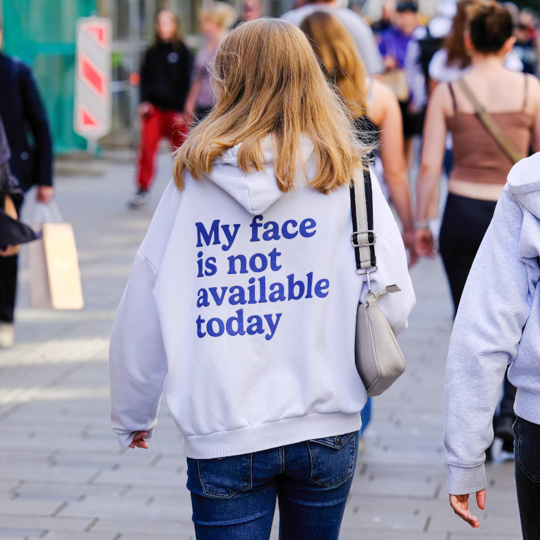 Eine Person geht eine Einkaufsstraße entlang, sie ist von hinten zu sehen. Sie trägt einen Pullover mit der Aufschrift auf dem Rücken: "My face is not availabel today."