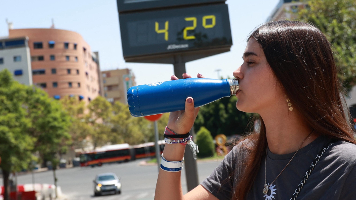 Eine junge Frau trinkt Wasser aus einer Flasche in Sevilla, Spanien. Im Hintergrund ist eine große Temperatur-Anzeige zu sehen, die 42 Grad Celsius anzeigt.