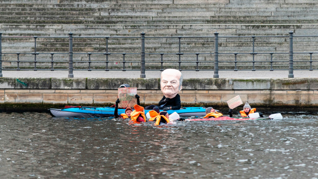 Ein Schauspieler mit einer großen Olaf-Scholz-Maske sitzt auf einem ausblasbaren Kanu auf der Spree. Um ih herum schwimmen Personen in orangenen Rettungswesten.