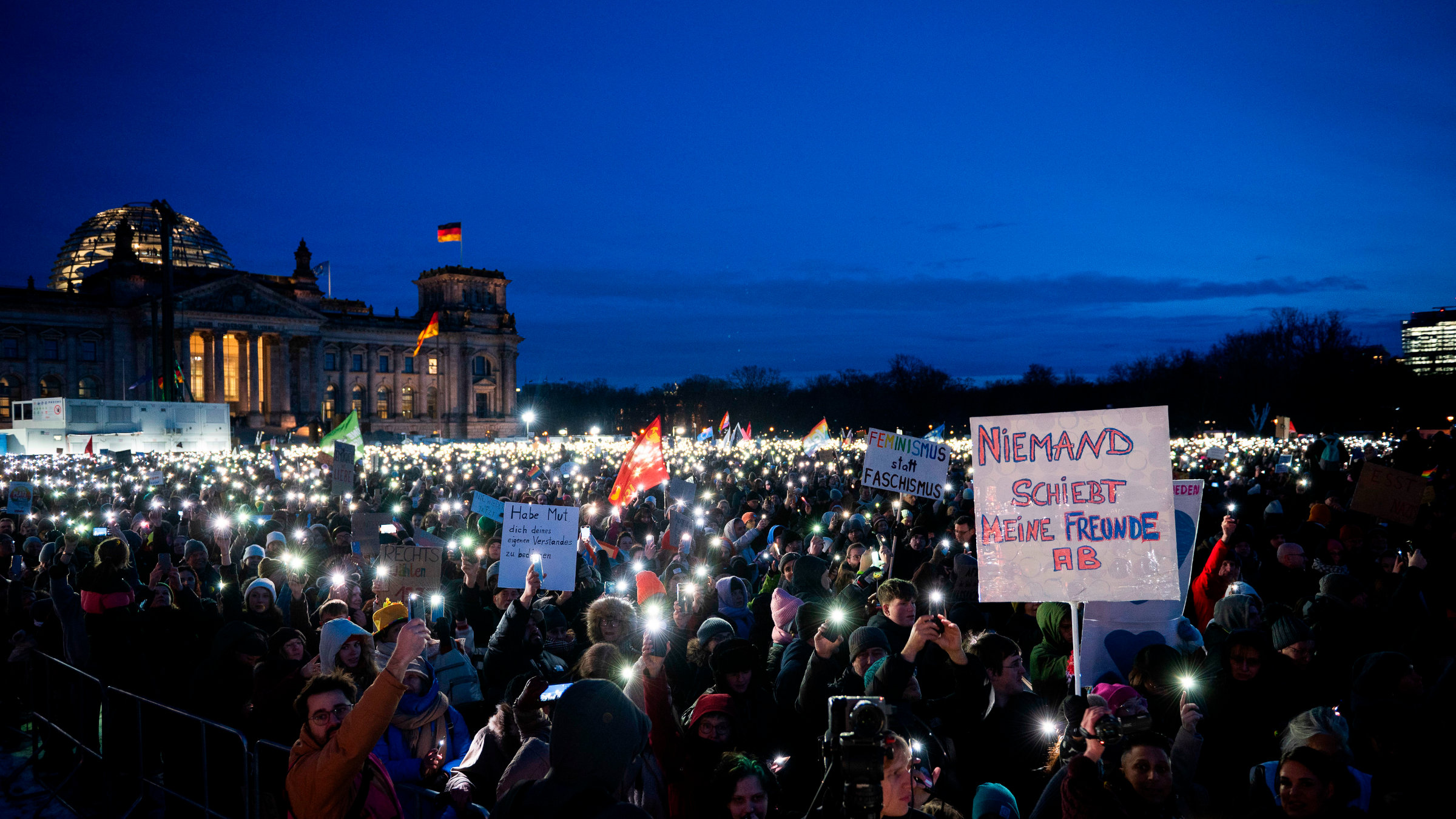 Eine große Menschenmenge versammelt sich nachts vor dem Reichstagsgebäude in Berlin, hält beleuchtete Handys und Protestschilder hoch, mit Botschaften wie „Feminismus statt Faschismus“ und „Niemand schiebt meine Freunde ab.“